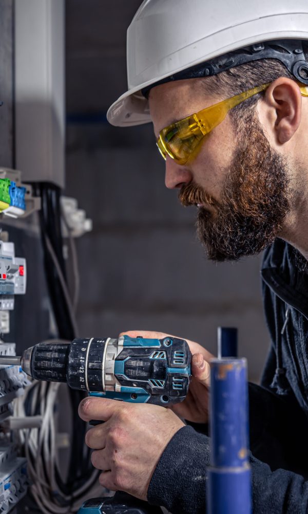 A male electrician works in a switchboard with an electrical connecting cable, connects the equipment with tools.